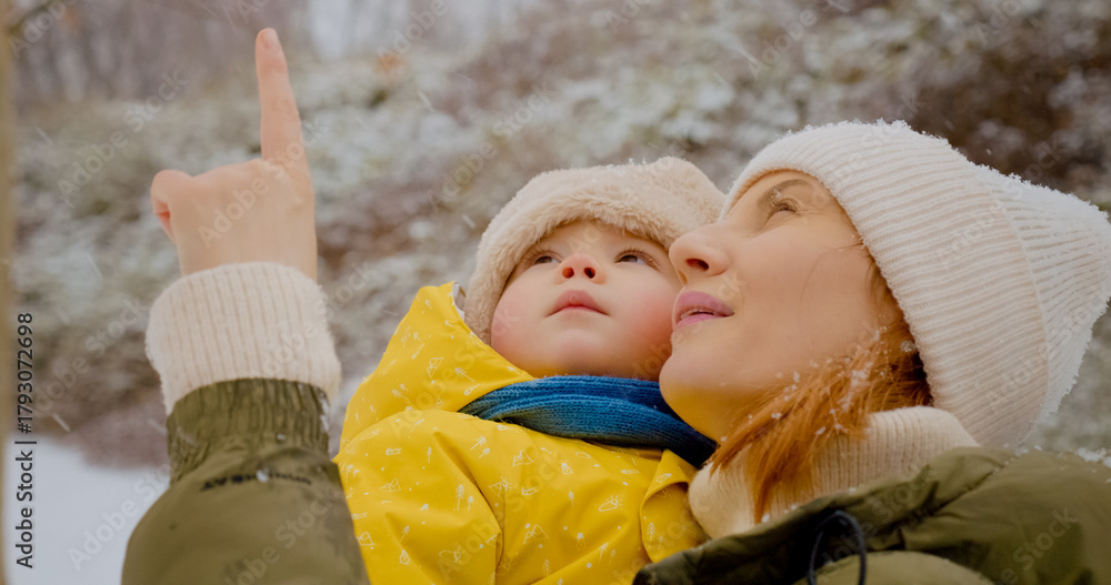 Obraz premium A mother holds her little boy while they delight in the snowfall at a park. They share joyful moments surrounded by winter scenery and playful snowflakes.