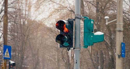 Fototapeta Naklejka Na Ścianę i Meble -  Snow continuously blankets the city as it gently falls. A red traffic light flickers above, directing the flow of vehicles while winter paints the landscape white.