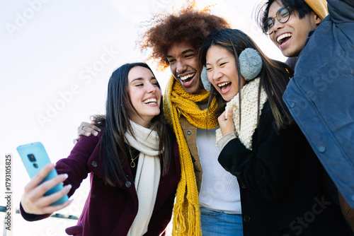 Diverse group of teenage friends having fun taking selfie portrait at city street on winter. Holiday lifestyle and technology concept