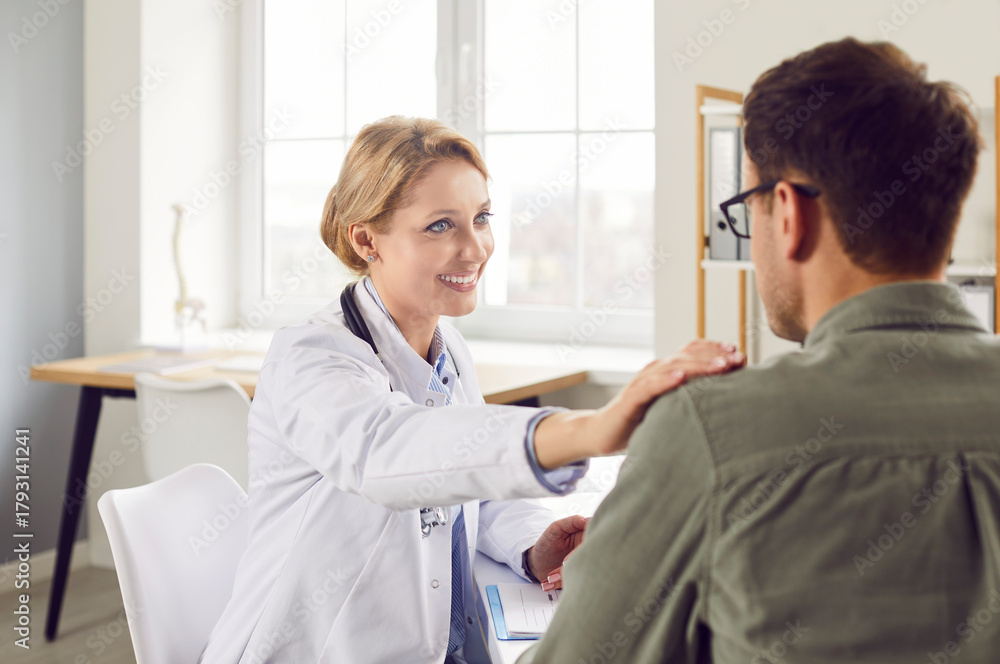 Obraz premium Portrait of female blonde friendly doctor supporting young male patient putting hand on his shoulder. Physician sitting in medical clinic giving consultation a man during medical exam in office.