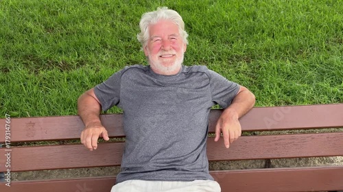 Happy cheerful elder 70s man sitting in a bench in the park, senior smiling bearded grandfather enjoying free time and nature in outdoors