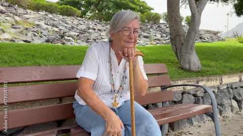 Senior elder 70s woman with walking disability holding knob of cane, sitting on a bench in the park to rest and relax, suffering from muscles problems, bones disease, recovering after trauma