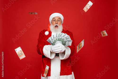Photo of pensioner old man grey beard raise fists open mouth money shower hold cash wear Santa Claus traditional costume red coat hat standing isolated red background