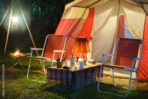 Camping tents at night, vintage cabin tents, antique oil lamps, retro chairs, and a group of camping tents with outdoor coffee-making facilities on wooden tables in a forest camping area.