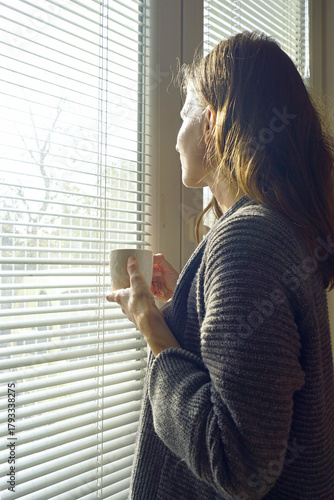 Billede på lærred Woman holding a coffee mug and gazing through window blinds as morning sunlight illuminates her profile