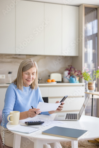 Smiling senior woman managing finances, calculating expenses, and paying bills using a laptop, calculator, and smartphone at the kitchen table, representing financial planning and retirement
