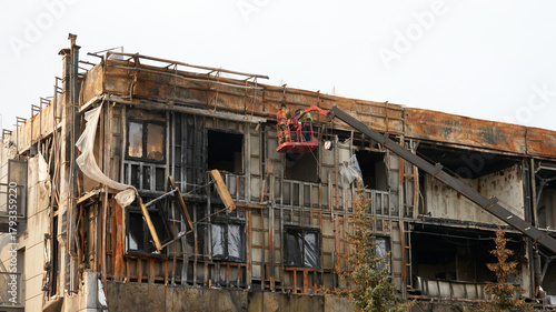 Experts in a construction cradle are assessing the damage to the building from an internal fire. The façade is severely burned, covered in soot, and structural elements are damaged.