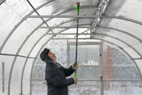 Protecting a polycarbonate greenhouse from deformation caused by snow accumulation on the roof. A man inside the greenhouse taps the dome with a special stick to remove the ice crust that has formed.