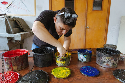 Person mixing yellow ink into a container, surrounded by colorful ink cans on a wooden table, preparing for printmaking