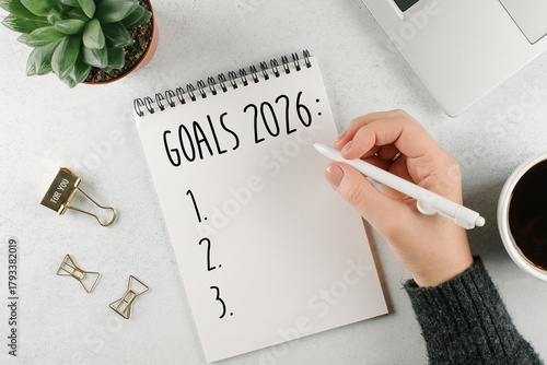 Woman's hand writing 2025 Goals in notebook. Laptop, home plant, cup of coffee on light table. Top view, flat lay