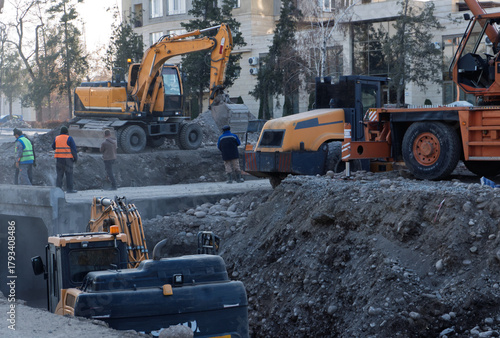 Several construction vehicles and workers are present at an urban construction site. Excavators and rollers are operating near a deep trench with visible dirt and debris.