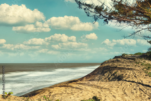 Sandy dune with ocean waves under a cloudy blue sky
