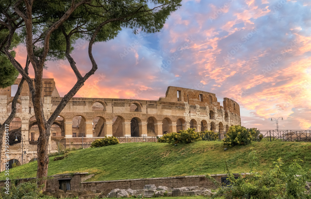 Obraz premium Rome, Italy. Ancient Colosseum (Coliseum) stands majestically against a vibrant sunset sky, surrounded by lush greenery and trees, showcasing historical architecture and cultural significance