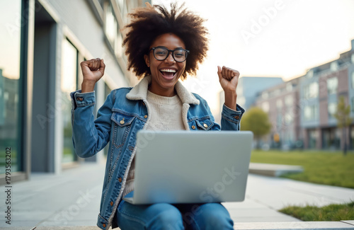 Excited african student with afro hair and glasses celebrates success. Girl student sits with laptop computer outside and smiles, clenching fists in winner gesture. Triumph concept and victory online.