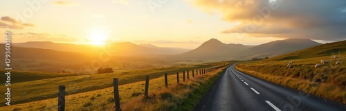 Rural road landscape at sunset. Winding road through green fields towards mountains. Sheep graze at the side of the road. Beautiful scenery during golden hour.