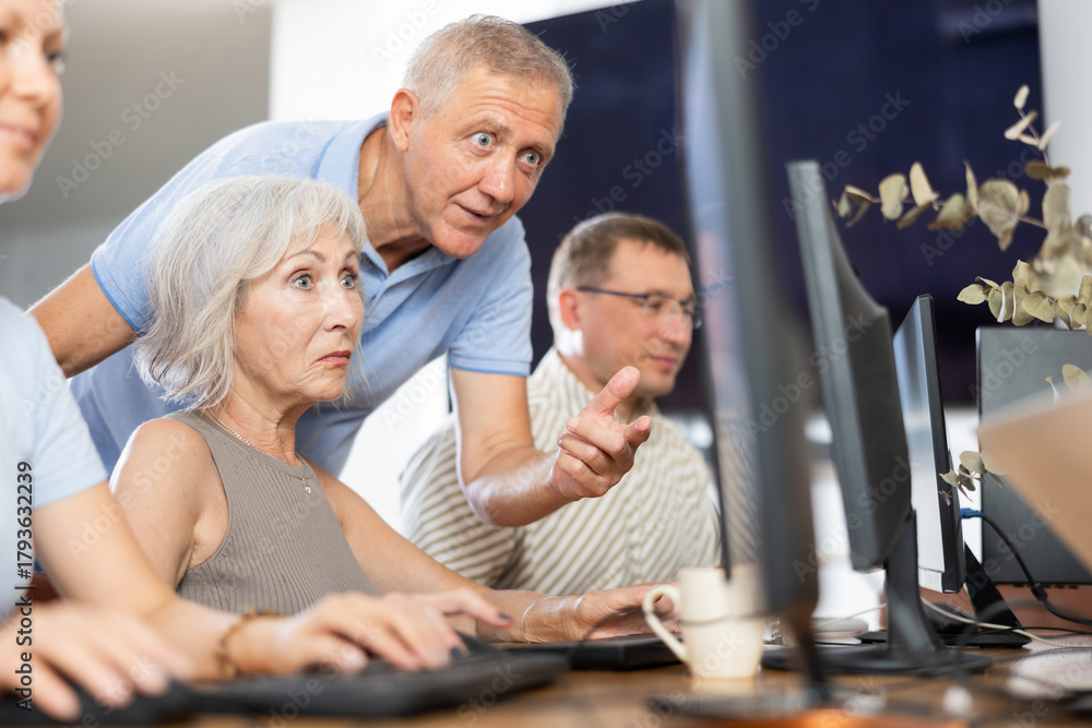 Fototapeta premium Smiling friendly older mentor explaining something to motivated elderly woman mastering computer skills during group course for adults..