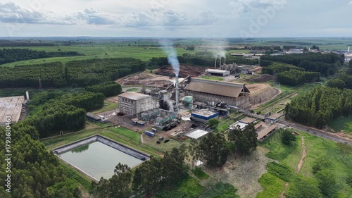 Aerial view of an industrial plant emitting steam into the atmosphere, set against a backdrop of trees and a pond