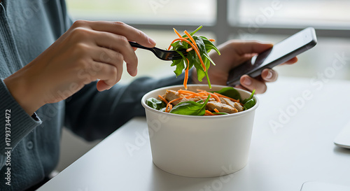 Woman eating a salad from a takeaway container while looking at her mobile phone.