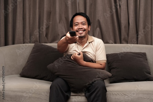 Southeast Asian man in a beige shirt sitting on a sofa, smiling while pointing a TV remote control forward in a relaxed indoor setting.
