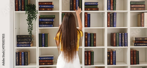 Female student taking book from shelving unit while studying at library, back...