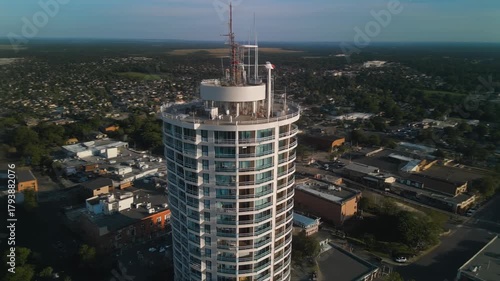 Aerial view of a modern cylindrical high-rise building with antennas on its roof, surrounded by a suburban landscape under a clear sky.