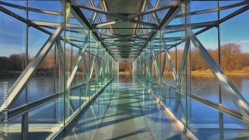 A modern glass and steel pedestrian bridge extends over calm water, offering a transparent pathway with reflections and a clear view of the distant trees.