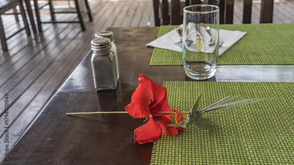 Fototapeta premium A bright red hibiscus flower and a bird woven from palm leaves lie on a table in a restaurant. There are napkins, a glass, cutlery, and spices on the countertop. Cuba