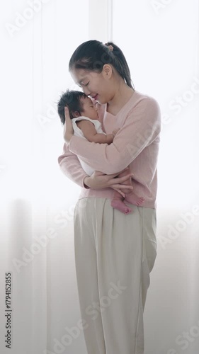 Asian mother standing full body view near curtain, holding newborn baby boy in arms while gently kissing, soft morning light, peaceful bonding moment between parent and infant