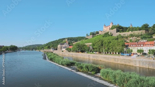 Wide-angle landscape of the Main river flowing past the Marienberg Fortress perched on a hill, overlooking the historic city and vineyards. Sunny day scene of German travel and architecture.