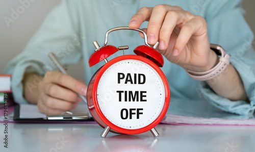 A person is writing on a piece of paper next to a red alarm clock that says Paid Time Off