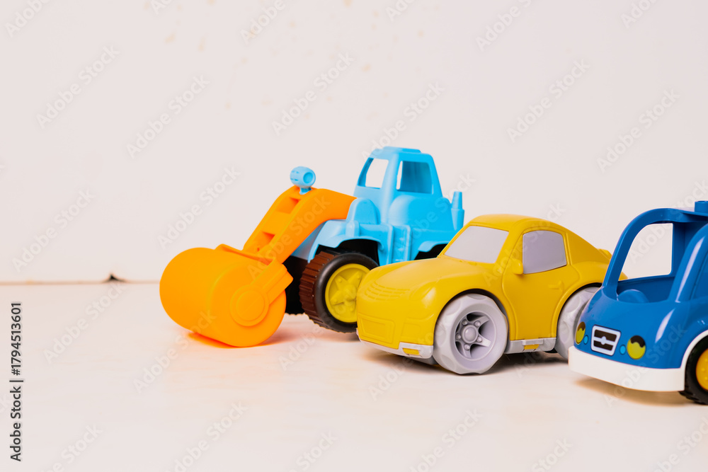 Obraz premium Four colorful toy vehicles are arranged in a row on a light wooden surface. The toys include a yellow truck, a blue car, a green vehicle, and an orange construction machine.