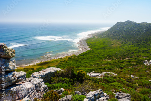 Scenic view from Gifkommetjie View Point, Cape Point, Table Mountain National Park. Rugged rocks and green fynbos overlook the Atlantic Ocean under clear blue skies in Cape Town, South Africa.