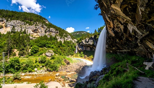 Waterfall cascades over rocks, creating a rainbow mist in a mountain landscape on a bright sunny day