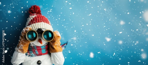 A snowman wearing a red knitted hat and scarf holds binoculars. Snowflakes fall against a blue background, creating a winter scene.