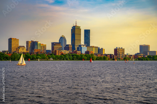 Beautiful sunset view over Boston, Massachusetts, featuring the iconic Back Bay skyline reflected on the Charles River. Small sailboats add a lively touch to the tranquil urban waterfront scene.