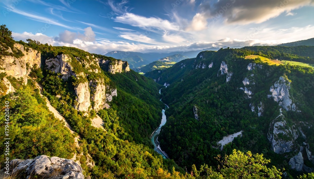 Fototapeta premium Winding river carves through lush green valley under a bright, partly cloudy sky, with distant mountains