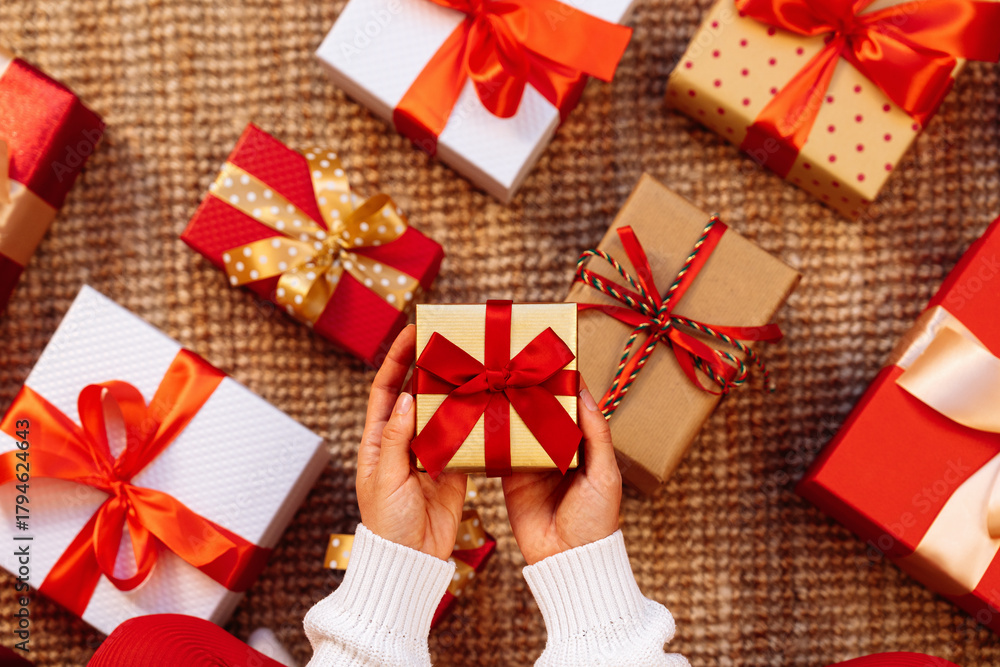 Obraz premium Small present with red ribbon and bow in hands above the gift boxes on the floor.