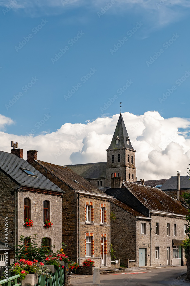 Obraz premium Traditional stone houses with flower boxes and a church tower in the historic village near Château de Spontin in Belgium showcasing authentic architecture and rural European charm under a vivid sky.