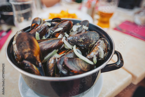 Freshly cooked mussels served in a black pot with sliced onions on a table at a seaside restaurant during sunset