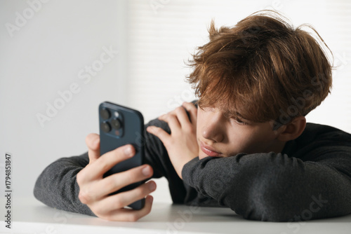 Sad teenage boy with smartphone at white table indoors
