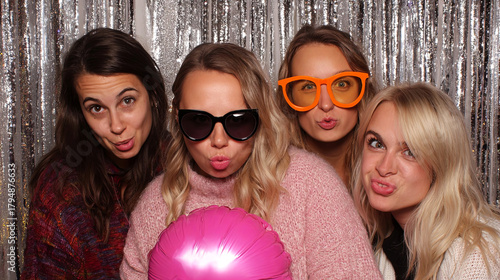 Group of four women posing playfully in a photo booth with colorful props, wearing oversized sunglasses and making funny faces, capturing a joyful moment of friendship