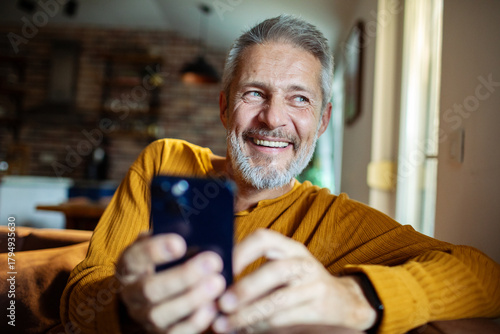 Картината върху платно Mature man happy and smiling while using smartphone at home
