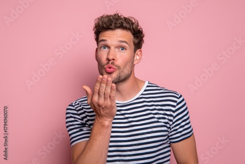 Attractive young man in a striped tee against a pink backdrop blowing a kiss and radiating charm