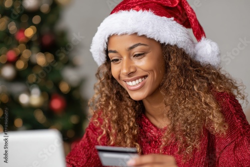 A cheerful young Black woman wearing a Santa hat purchases gifts online with a credit card and laptop