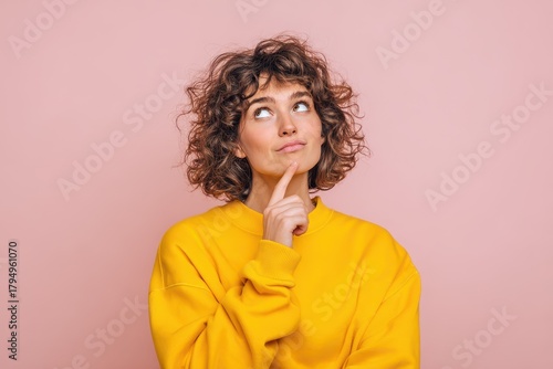 A cheerful young woman in a yellow sweatshirt rests her chin on her hand deep in thought set against a soft pink background