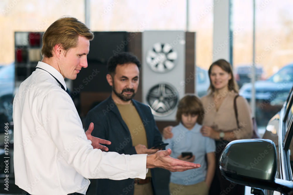 Fototapeta premium Caucasian middle aged man gesturing while explaining car features to Middle Eastern middle aged man with beard and Caucasian woman and child