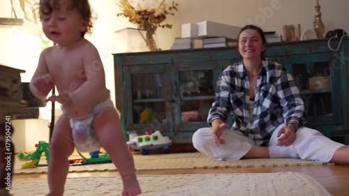 Toddler taking first independent steps while trying to keep balance, stumbling but determined to stand again as mother encourages with pride and joy in background