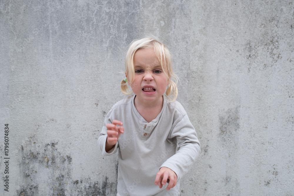 Fototapeta premium Little 4 Year Old Blonde Girl Standing by a Wall, Pretending to Be Angry and Showing Claws