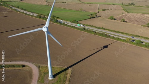 Aerial view of a towering wind turbine casting a long shadow over the brown fields, near a highway with passing vehicles, Melton Mowbray, England, United Kingdom.