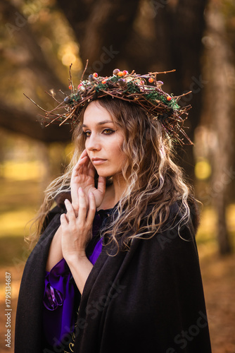 A young woman stands in a golden autumn forest, wearing a black medieval dress with purple accents. She wears a crown of twigs and berries, and a dark hooded cloak. This fantasy portrait  